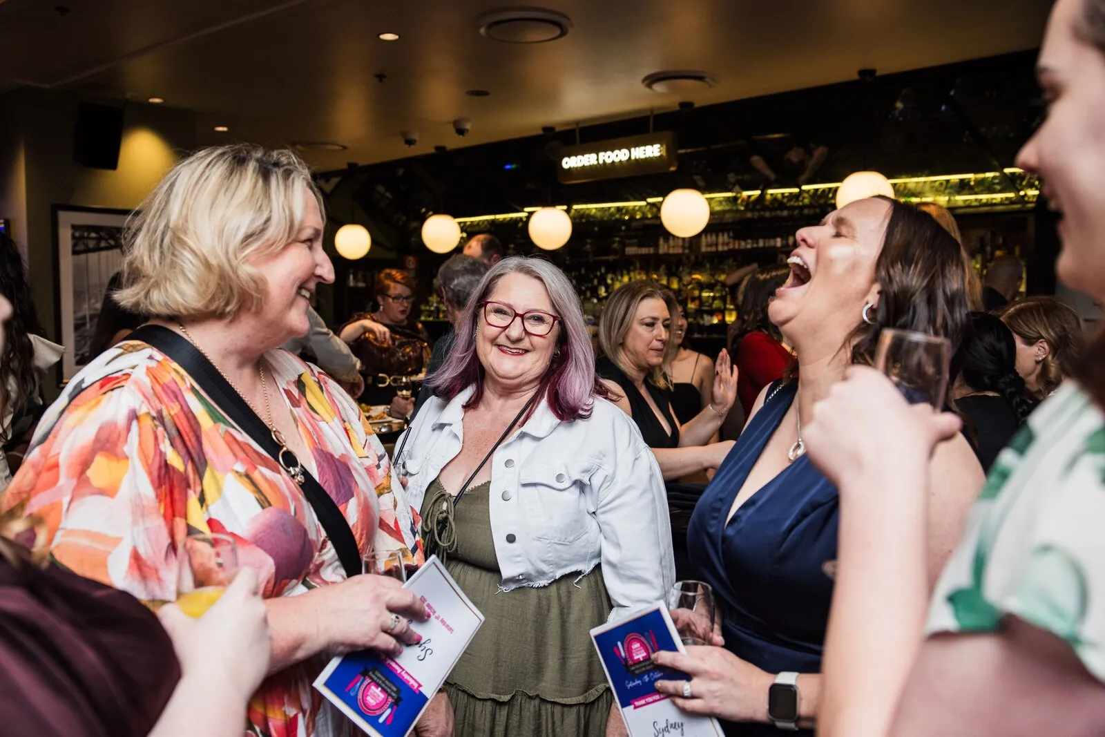 A lively group of guests laughing together at the bar reception, drinks and programs in hand