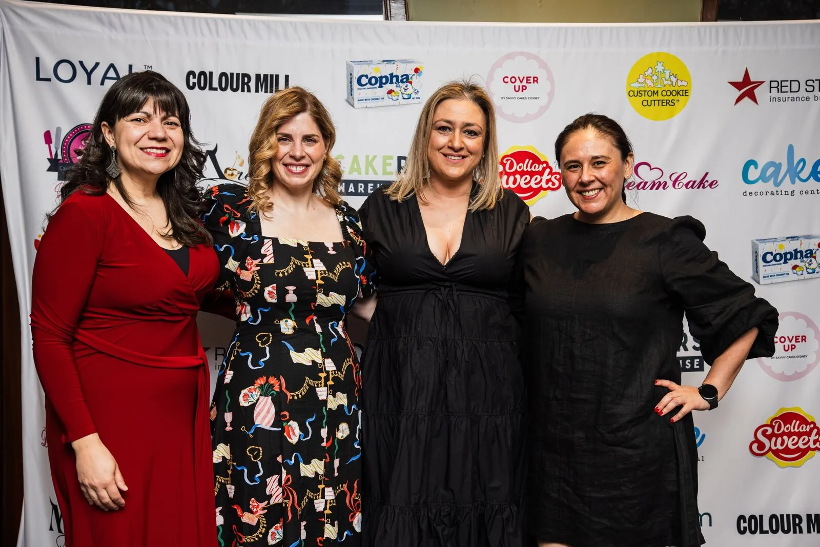 Four women in eveningwear posing together in front of the sponsor banner