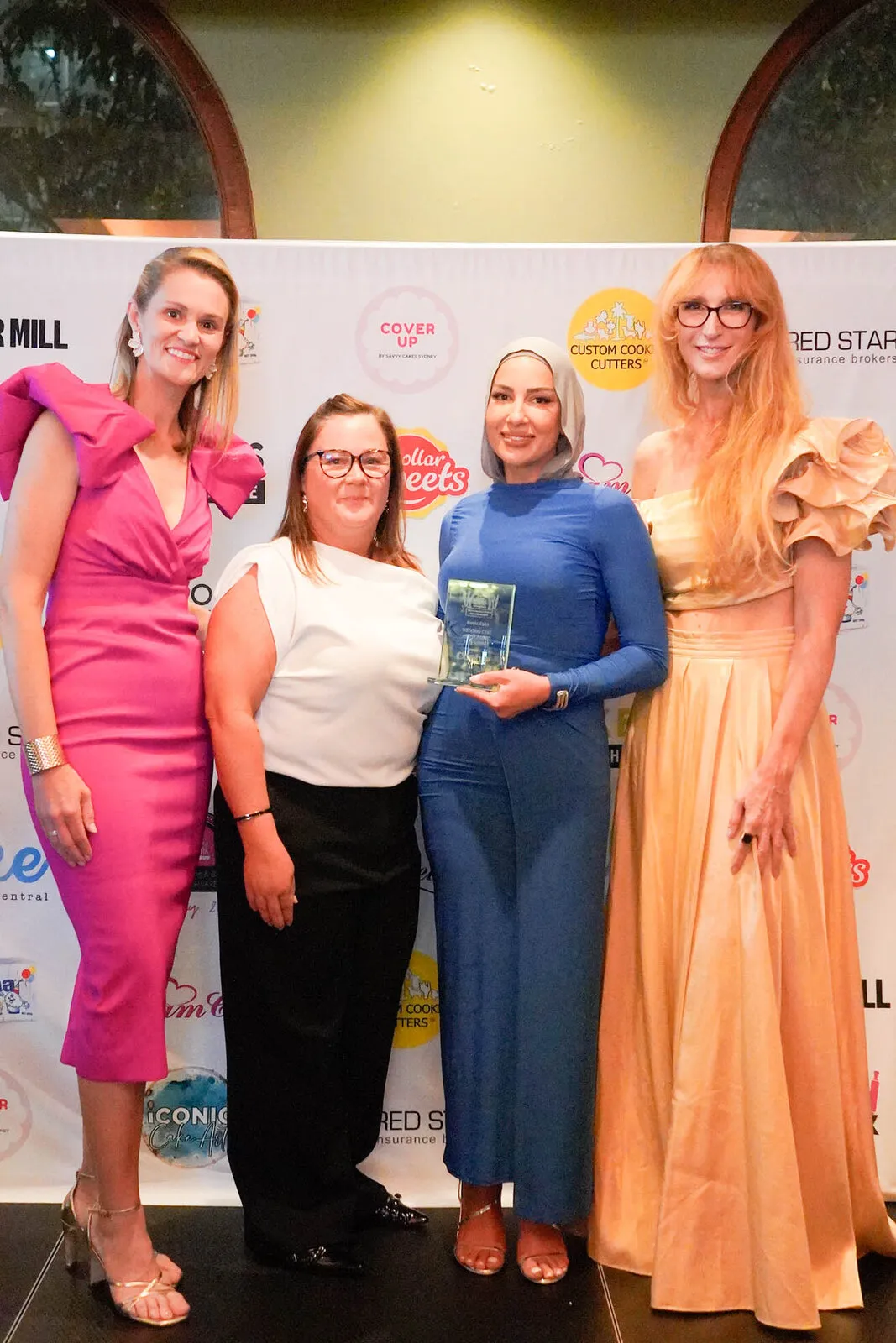 Four women in eveningwear in front of the sponsor banner, one holding a glass trophy