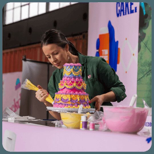 A decorator in a green apron piping colourful scalloped petals onto a tall tiered cake during a live demonstration at a Cake! event