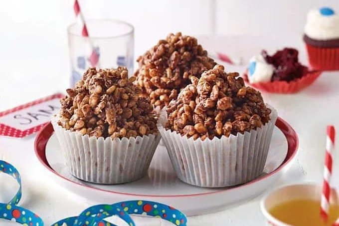 Three chocolate crackles in white patty cases on a red-rimmed plate, with party ribbon and a paper cup beside them.