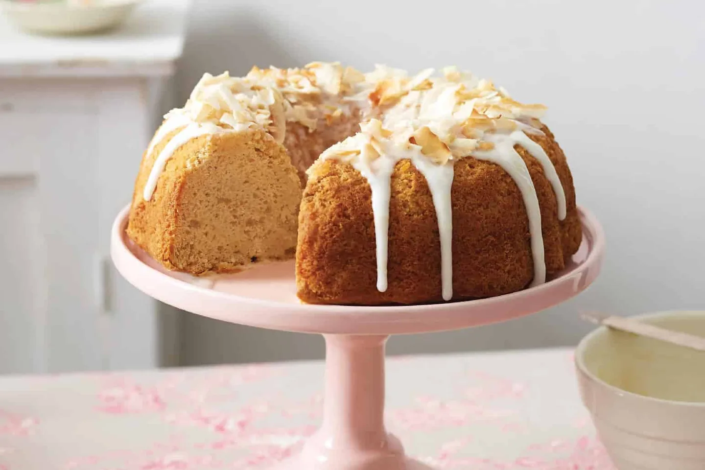 A banana cake iced and topped with toasted coconut on a pink cake stand, with one slice cut and plated.